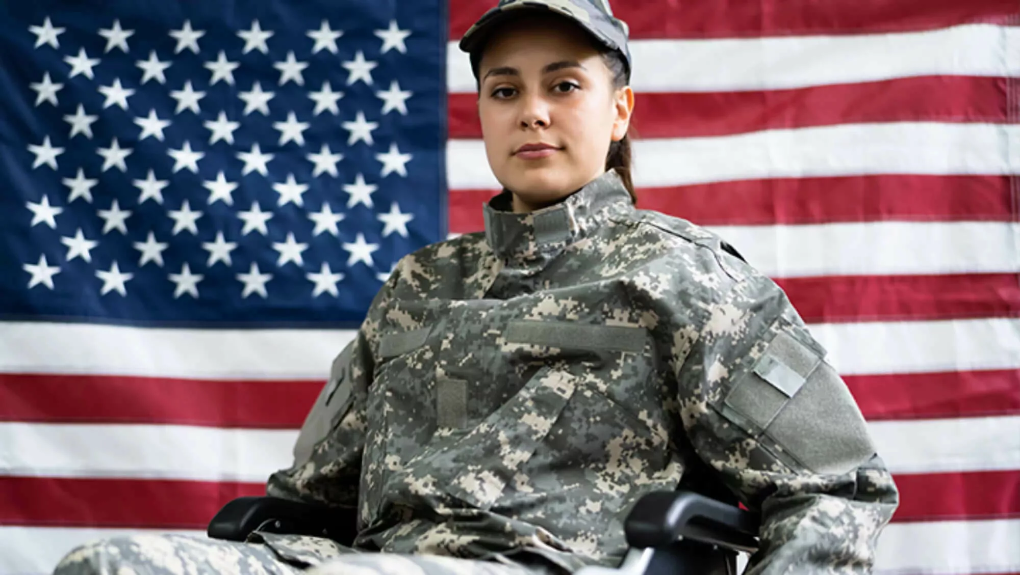Female veteran in uniform in a wheelchair in front of the American flag.
