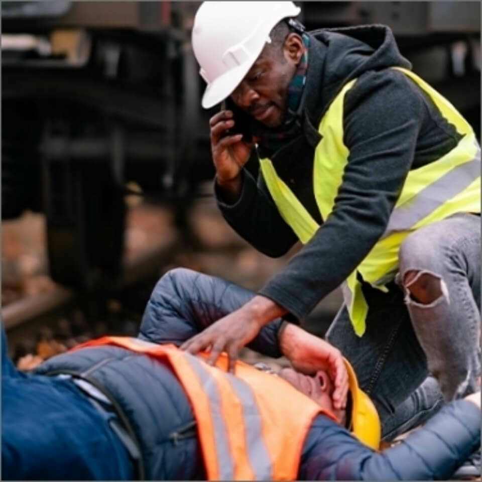 Fallen construction worker with another worker leaning over him while making a phone call.