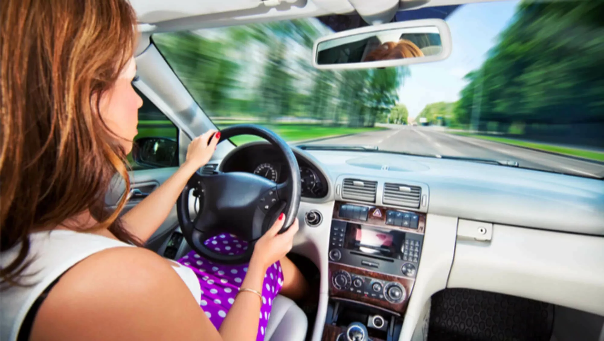 Woman in purple polka dots behind the wheel of a car, driving quickly.