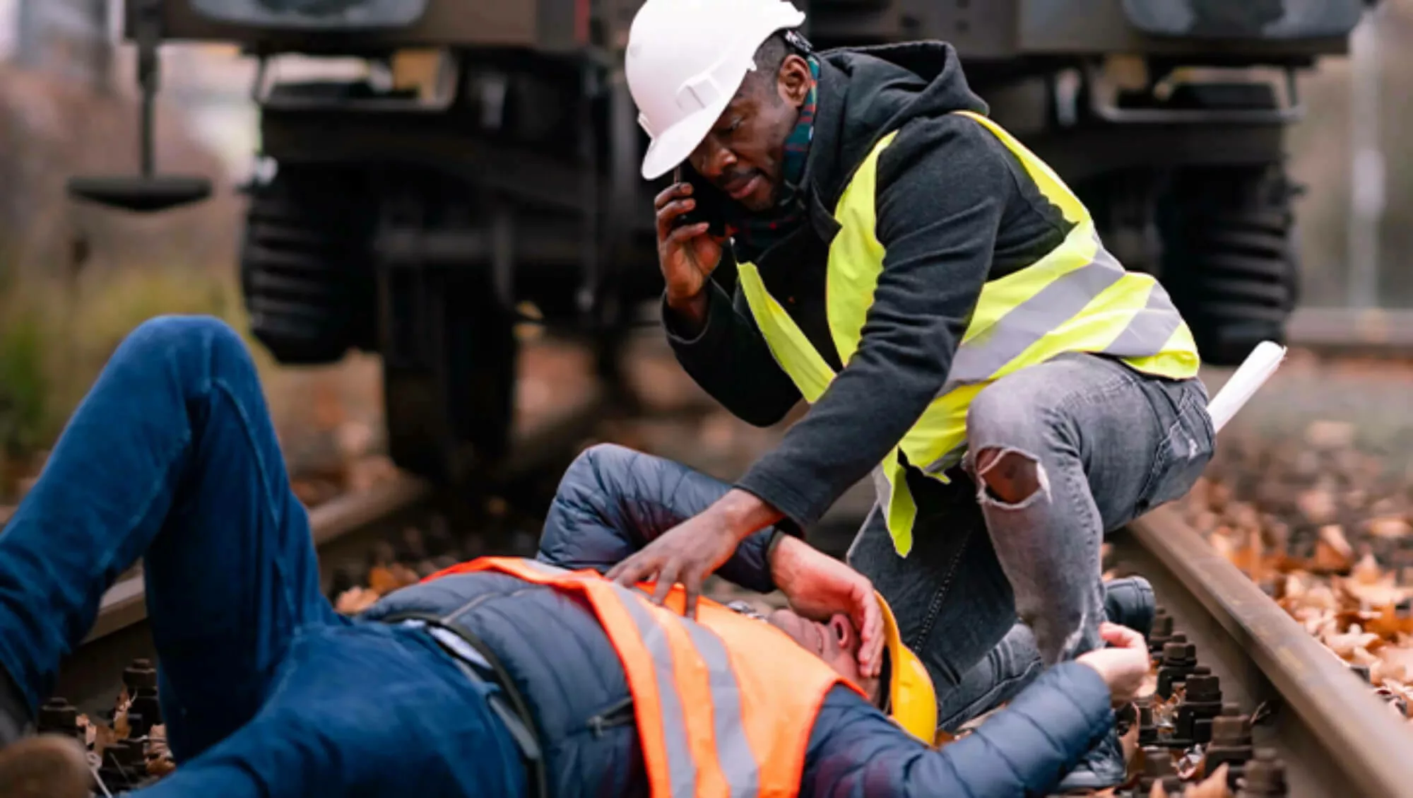 Fallen construction worker with another worker leaning over him while making a phone call.