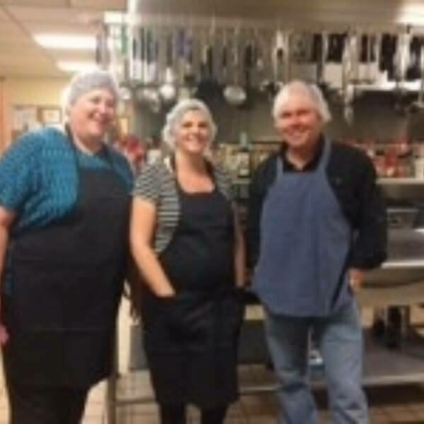 Three people standing in a kitchen in aprons and hair nets.